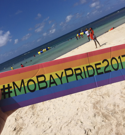 Montego Bay Pride 2017 hashtag (#MoBayPride2017) on a sign held in hand with the beach in Montego Bay, Jamaica, in the background.