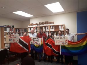 Six individuals standing in a row, holding the flags of Trinidad and Tobago and the rainbow flag.